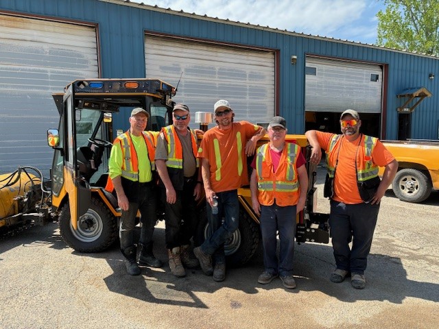 public works crew stands in front of machinery and shop